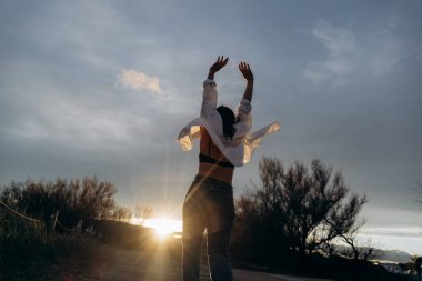 Kadın, kollarını gökyüzüne kaldırmış, güneş doğanın üzerinde batarken özgürlüğünü ve neşesini ifade ediyor. Açık tişört ve günlük kıyafet tasasız bir anı ifade eder..