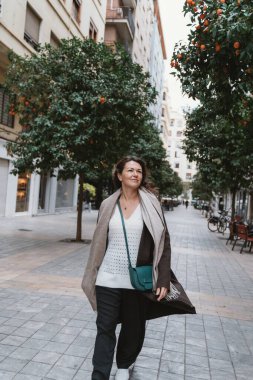 Bir kadın turuncu ağaçlarla kaplı bir caddede yürüyor, günlük kıyafetler giyiyor ve çapraz ceset torbası taşıyor. İfadesi sakin ve sakin, şehir ortamında..