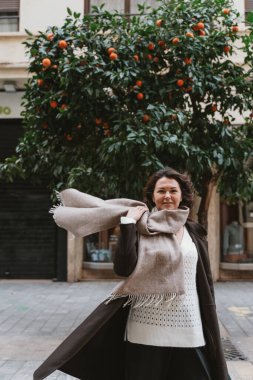 Bir kadın, şehir caddesinde yemyeşil bir portakal ağacının önünde kendine güvenen büyük bir atkıyla ayakta duruyor. Sahne kentsel ve doğa öğelerinin bir karışımını yakalıyor..