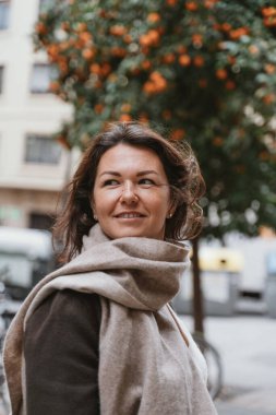 Woman in cozy scarf smiling outdoors with orange trees and buildings in the background. Hair gently blown by the wind, creating a serene and confident expression.