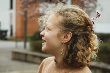 Neşeli kız saçlarında kiraz çiçeği dallarıyla gülüyor, mutluluk ve tarz saçıyor. Yan profil, gençlik enerjisini ve çiçeklerle süslenmiş neşeli bir anı yakalıyor..