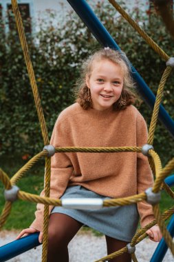 Çocuk neşeli bir ifadeyle oyun parkında ip üzerinde oturuyor. Bir süveter ve etek giyerek, çevresi yeşillik ve oyun ekipmanlarıyla çevrili bir gün geçirmekten hoşlanıyor..