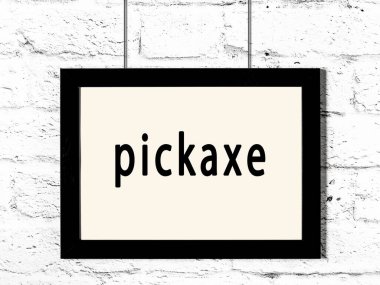 Siyah ahşap çerçeve, beyaz tuğla duvara kazma asılmış. 