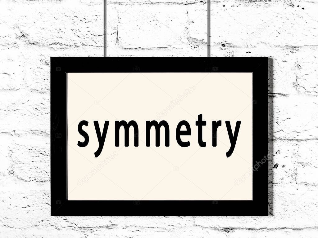 Black wooden frame with inscription symmetry hanging on white brick wall 