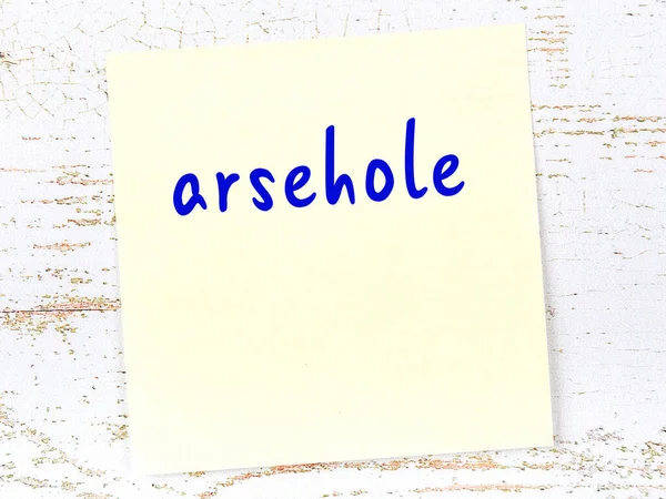 Yellow sticky note on wooden wall with handwritten inscription arsehole