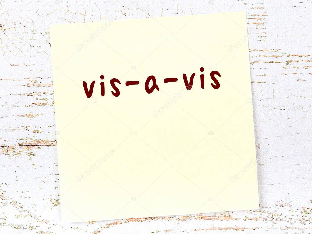 Yellow sticky note on wooden wall with handwritten inscription vis-a-vis