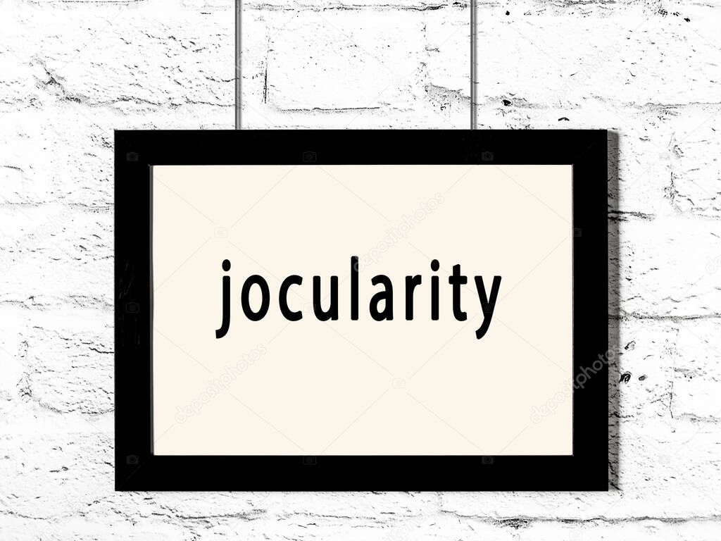 Black wooden frame with inscription jocularity hanging on white brick wall 