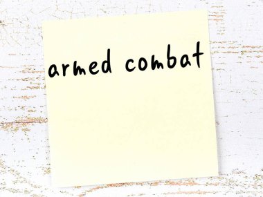 Tahta duvardaki sarı yapışkan notta el yazısıyla yazılmış silahlı çatışma yazısı var.