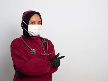 Beyaz arka planda izole edilmiş bir maske takan genç bayan doktor şaşırır ve parmağını yukarı doğru gösterir. Asyalı Müslüman hemşire kırmızı bir ameliyat elbisesi giyiyor ve parmağıyla yan tarafı gösteriyor..