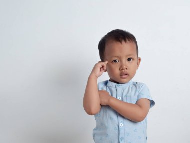 Asyalı bir çocuk düşünme jestinde poz veriyor. Yeni yürümeye başlayan çocuk, beyaz bir arka plan karşısında düşünme işareti olarak işaret parmağıyla alnının yanına poz veriyor..