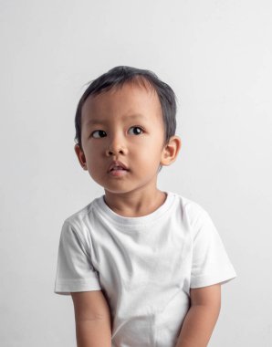 Sakin bir Asyalı çocuk, düşünceli bir ifadeyle oturur, beyaz bir tişört giyer, düz bir arka plana yaslanır, çocuk portresi, duygu veya eğitim konsepti için uygundur..