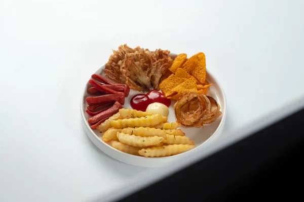 Çıtır soğan halkaları, patates kızartması, tortilla cipsi, kızarmış mantar, ketçaplı sosis ve mayonez sosisi de dahil olmak üzere beyaz bir tabakta çeşitli kızarmış atıştırmalıklar..
