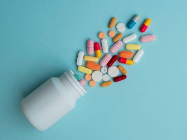 Beyaz kavanoz plastiğinden mavi arka plana çeşitli renkli antibiyotik kapsülleri dökülüyor. Çeşitli renkli reçeteli kapsül hapları. Sağlık hizmetleri, ilaç tedavileri ve ilaç endüstrisi için idealdir..