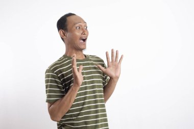 Asyalı bir adam yeşil polo tişörtü giyiyor, etkileyici ve ürkek bir jest yapıyor, ellerini kaldırıyor, sanki şokta ya da savunmada gibi, düz beyaz bir arka plana karşı duruyor,