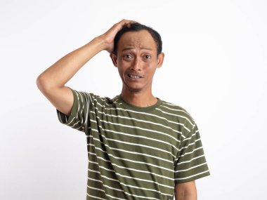 Yeşil gömlek giyen Asyalı bir adam, düz beyaz bir arka plana karşı duruyor, iki eliyle başını tutuyor ve acı verici ve stresli bir yüz ifadesi sergiliyor, baş ağrısı tasvir ediyor.