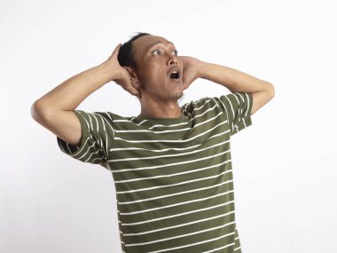 Yeşil t-shirt giyen Asyalı bir adam, düz beyaz bir arka planın önünde duruyor, başını iki eliyle tutuyor ve şaşkınlık ve şaşkınlık dolu bir yüz ifadesi sergiliyor, bir şeyi inkar ettiğini gösteriyor..