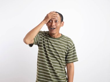 Yeşil gömlek giyen Asyalı bir adam, düz beyaz bir arka plana karşı duruyor, elleriyle başını tutuyor ve acı verici ve stresli bir yüz ifadesi sergiliyor, baş ağrısı tasvir ediyor.