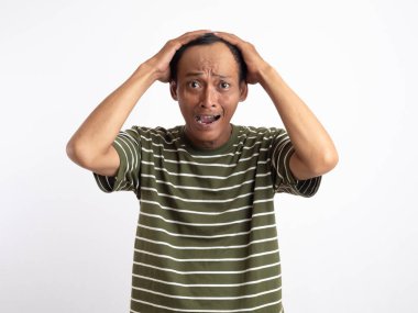 Yeşil t-shirt giyen Asyalı bir adam, düz beyaz bir arka planın önünde duruyor, başını iki eliyle tutuyor ve şaşkınlık ve şaşkınlık dolu bir yüz ifadesi sergiliyor, bir şeyi inkar ettiğini gösteriyor..