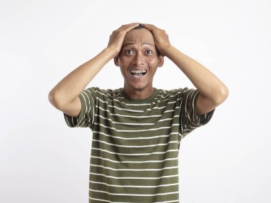 Yeşil gömlek giyen Asyalı bir adam, düz beyaz bir arka plana karşı duruyor, elleriyle başını tutuyor ve acı verici ve stresli bir yüz ifadesi sergiliyor, baş ağrısı tasvir ediyor.