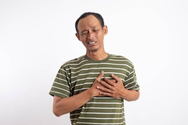 Yeşil polo tişörtlü Asyalı bir adam göğsünü iki eliyle kavrar, acı ya da duygusal sıkıntı ifade eder, öne doğru hafifçe eğilir, dramatik bir şekilde beyaz bir arkaplana yaslanır.