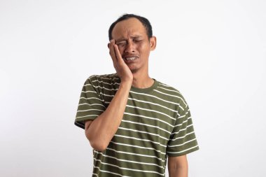 Beyaz çizgili yeşil bir gömlek giyen bir adam, bir eliyle yanağını tutarken diğer eliyle karnını tutuyor, düz bir arka planda duruyor, acıyı ifade ediyor.