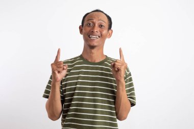 Yeşil gömlekli Asyalı bir adam her iki işaret parmağını da yukarı kaldırıyor, beyaz bir arka plana doğru gülümsüyor, bir fikri vurguluyor ya da dikkatleri başka bir şeye çekiyor.