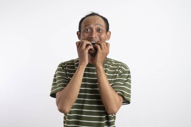 Tişörtlü, ağzı açık, gözleri açık, parmaklarını ısıran, mavi stüdyo arka planında izole edilmiş Asyalı bir adamı şok etti. Kapatın. Korku, insani duygu, şaşkınlık, yüz ifadesi