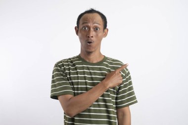 Yeşil tişörtlü Asyalı bir adam, sanki bir reklam aracı için beklenmedik bir şey fark ediyormuş gibi, şaşırmış ve meraklı bir ifadeyle yan tarafı işaret ediyor. Beyaz arka plan onun heyecanlı ifadesini vurguluyor..