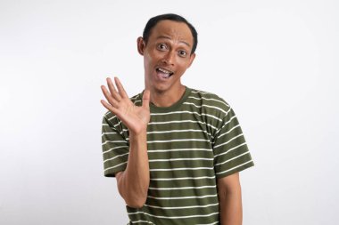 Beyaz çizgili yeşil gömlek giyen Asyalı bir adam geniş ağızlı bir gülümseme ile coşkuyla dalgalanır, açık gri bir stüdyo arka planına karşı durur. Onun neşeli ifadesi heyecan ve dostluğu iletir.