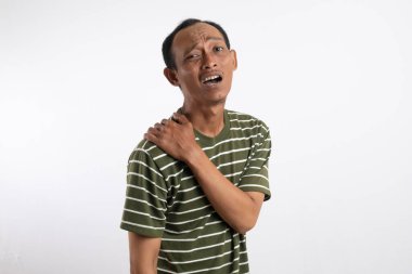 Beyaz çizgili yeşil gömlek giyen bir adam omzunu acılı bir ifadeyle tutar, bu da omuz ağrısı ya da rahatsızlığı gösterir. Açık mavi arka plan sıkıntısını vurguluyor.