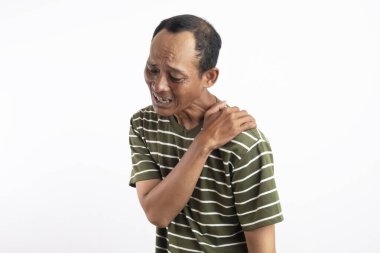 Beyaz çizgili yeşil gömlek giyen bir adam omzunu acılı bir ifadeyle tutar, bu da omuz ağrısı ya da rahatsızlığı gösterir. Açık mavi arka plan sıkıntısını vurguluyor.