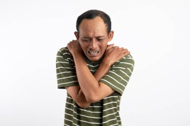 Beyaz çizgili yeşil gömlek giyen bir adam omzunu acılı bir ifadeyle tutar, bu da omuz ağrısı ya da rahatsızlığı gösterir. Açık mavi arka plan sıkıntısını vurguluyor.