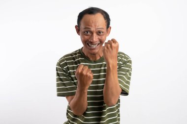 Beyaz çizgili yeşil gömlek giyen bir adam kutlama jesti olarak her iki elini de kaldırır, yüzü heyecan ve neşe içinde ışıldar. Açık mavi arka plan onun zafer ve coşkulu ifadesini vurguluyor..