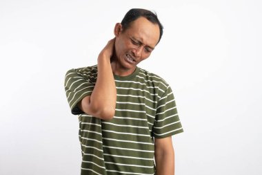 Yeşil gömlek giyen Asyalı bir adam, düz beyaz bir arka plana karşı duruyor, boynunu tutuyor ve rahatsızlık gösteriyor, boyun ağrısını, gerilimi veya gerilimi sıradan bir pozda resmediyor.