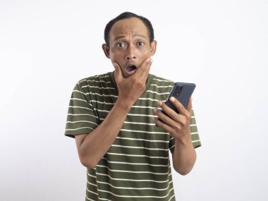 Yeşil tişörtlü genç Asyalı adam pembe arka planla izole edilmiş stresli bir ifadeyle akıllı telefonuna bakıyor..