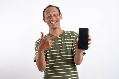 Yeşil gömlekli bir adam elinde boş bir ekranla akıllı telefonu tutarken gülümsüyor. Arka plan bembeyaz ve adam diğer eliyle kamerayı işaret ediyor.