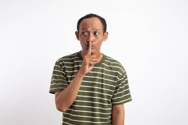 Beyaz çizgili yeşil gömlekli bir adam beyaz bir arka plana karşı duruyor, işaret parmağı dudaklarında sessizlik için işaret ediyor. İşaret parmağı ağzın önünde, sessizlik jesti