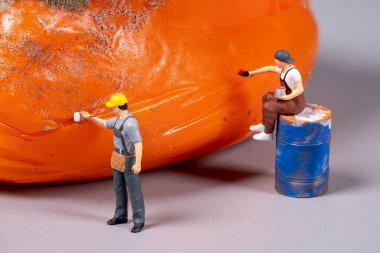 İki minyatür işçi büyük bir turuncu yüzeyi boyamakla meşguller. Biri boya sürerken ayakta duruyor, diğeri de bir fıçının üzerinde oturuyor..