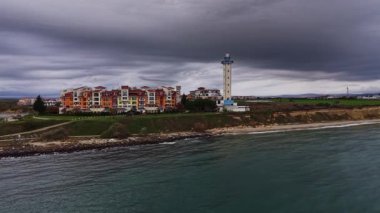 Bulgaristan 'da manzaralı bir kıyı şeridi yakalayan hava manzarasında, dramatik bulutlar ve sakin sulara karşı canlı binaların yanında bir deniz feneri sergileniyor..