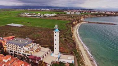 Bulgaristan 'da yeşil alanlar ve bulutlu bir gökyüzü altında sakin bir denize sahip bir deniz feneri ve yakındaki yapılara sahip bir kıyı manzarası hava manzarası sergiliyor.