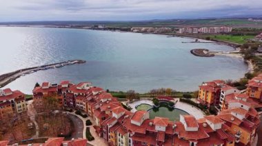 Sahne, Bulgaristan 'ın geniş bir kıyı bölgesini, kıyı boyunca uzanan renkli binaları, sakin suları ve kuş bakışı bir perspektiften uzak tepeleri gösteriyor..