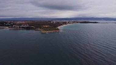 Bulgaristan 'ın kıyı şeridinin hava manzarası, öğleden sonra bulutlu bir gökyüzü altında binaların ve doğal özelliklerin karışımının yer aldığı manzaralı bir plaj sahası gösteriyor..