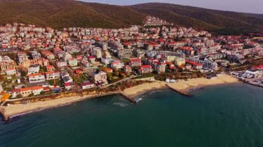 Bulgaristan 'ın sahil manzarasında, sahil boyunca renkli binalar ve arka planda verimli tepeler bulunan bir kıyı kenti ön plana çıkıyor. Sahne, deniz kenarında canlı bir topluluk yakalıyor..