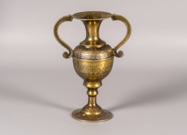 Tarafsız bir yüzeyde sapı olan altın bir vazo dikey duruyor. Vazonun ayrıntılı desenleri ve pürüzsüz bir kaplaması var..