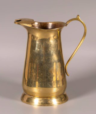 Pirinç bir sürahi nötr bir yüzeyde dimdik durur. İçinde kıvrımlı bir kulp ve bir çıkıntı var. Yüzey ışığı yansıtıyor, işçiliği ve stili gösteriyor..