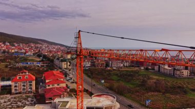 Uzun turuncu bir vinç gelişmekte olan bir bölgenin üzerinde duruyor ve kıyı kasabasını görüyor. Yerleşim yerleri ve deniz güneş batarken görünür..