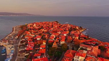 Bulgaristan 'ın Nesebar kentinin nefes kesici hava manzarası, çarpıcı bir günbatımında renkli çatılar ve kıyı şeridi sergiliyor. Su, turuncu ve pembenin ılık tonlarını yansıtır..