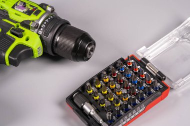 Bir matkap gri bir çalışma yüzeyindeki tornavida parçalarının yanında duruyor. Matkabın yeşil ve siyah bir tasarımı var. Parçalar plastik bir kutuda düzenlenir..