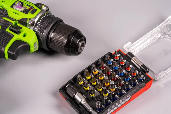 Bir matkap gri bir çalışma yüzeyindeki tornavida parçalarının yanında duruyor. Matkabın yeşil ve siyah bir tasarımı var. Parçalar plastik bir kutuda düzenlenir..