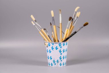 Bir kova, ahşap saplı ve metal ferruatlı çeşitli boya fırçaları taşır. Kovanın mavi bir deseni var ve gri bir yüzeyde duruyor. Çalışma alanındaki yaratıcılığı gösteriyor..